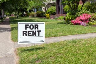 Sidewalk view of front yard of a property with black and white "For Rent" sign